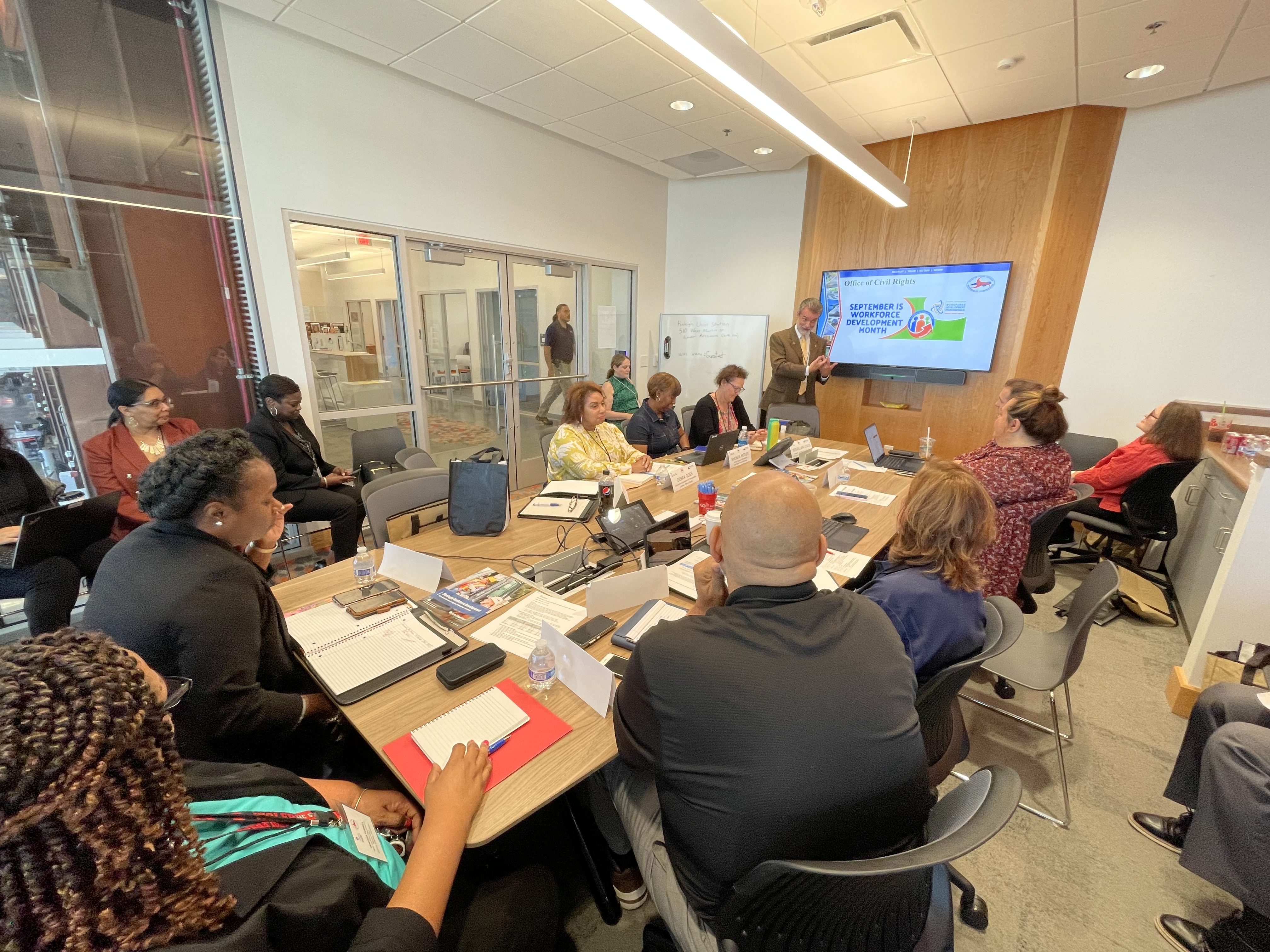 People gathered at a conference room table watching a man present something on a screen.